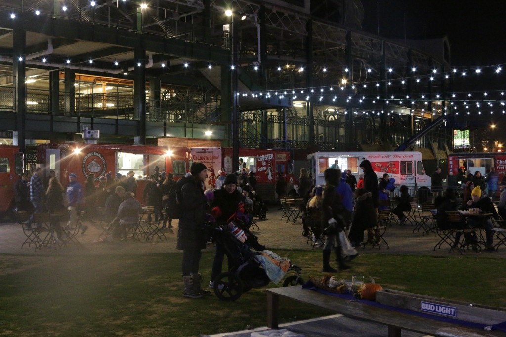 Crowd mingling near lit trees and a tall LED pillar beside Wrigley Field structures