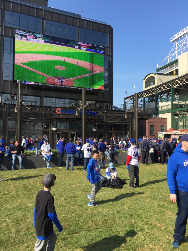 Fans on the grass near the Cubs Store and a large outdoor screen beside Wrigley Field