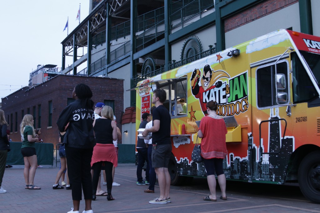 Crowd on the Gallagher Way lawn at twilight with food vendors and Wrigley Field behind them